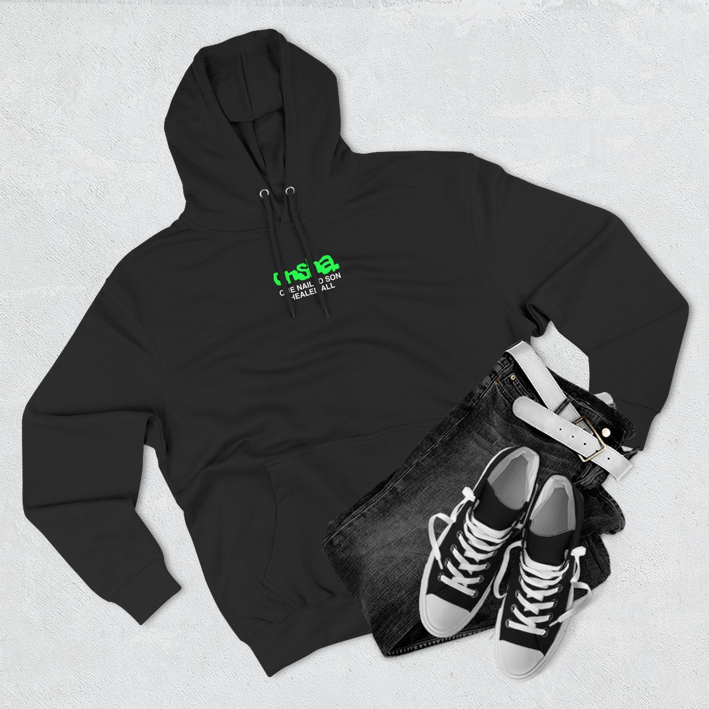 Flat lay of black Onsha hoodie with green chest logo 'One Nailed Son Healed All', black jeans and sneakers on light gray background